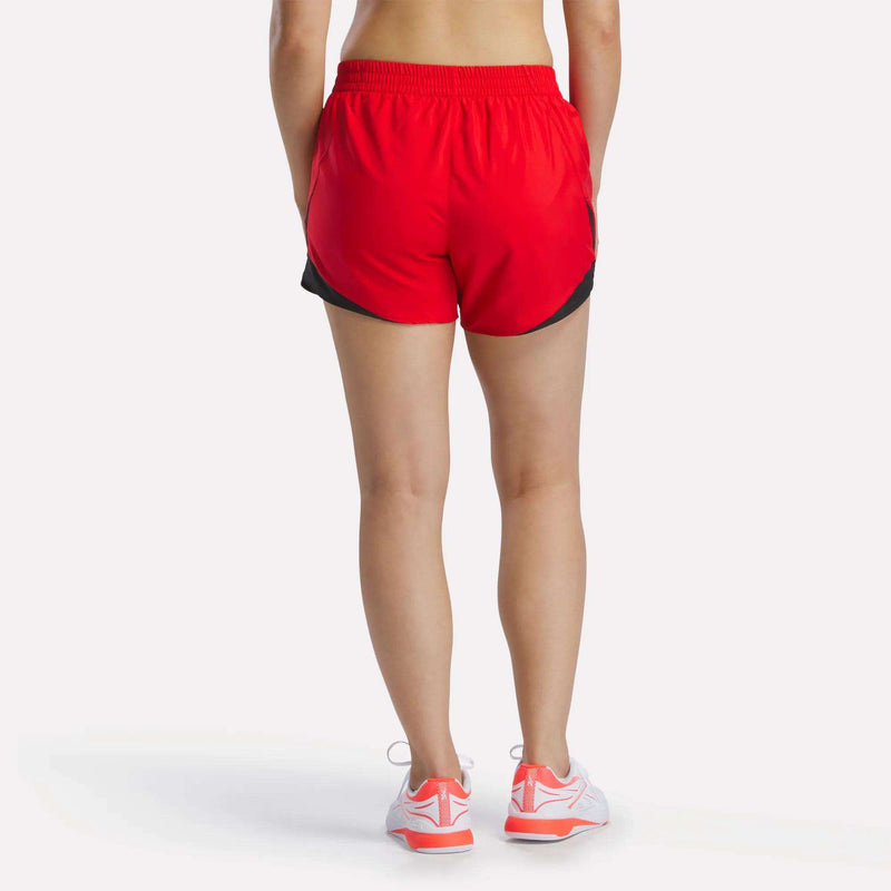 A person faces away wearing Reebok Women's Colorblocked Woven Shorts in bright red with black trim and an elastic waistband, paired with white socks and white running shoes with orange accents. The background is plain white.