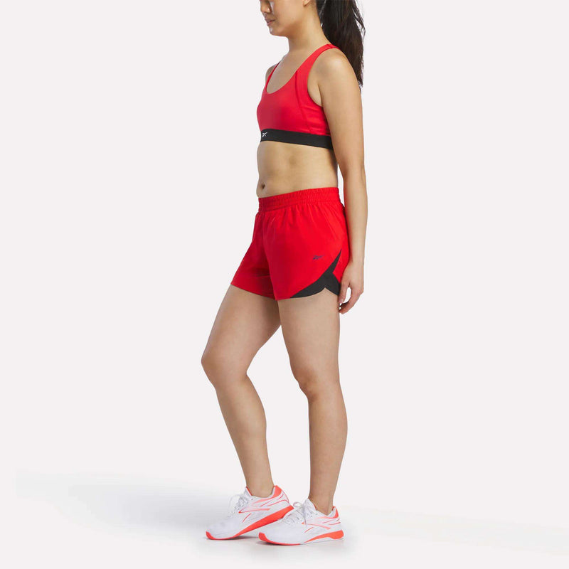A woman wearing a red sports bra and Reebok Women's Colorblocked Woven Shorts stands in profile against a white background, paired with white athletic shoes.