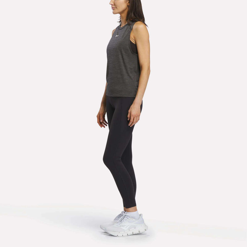A woman stands in profile against a plain white background, wearing the Reebok Women's Training Tank Top—a sleeveless gray design with RBK-DRY technology—paired with black leggings and white athletic shoes.