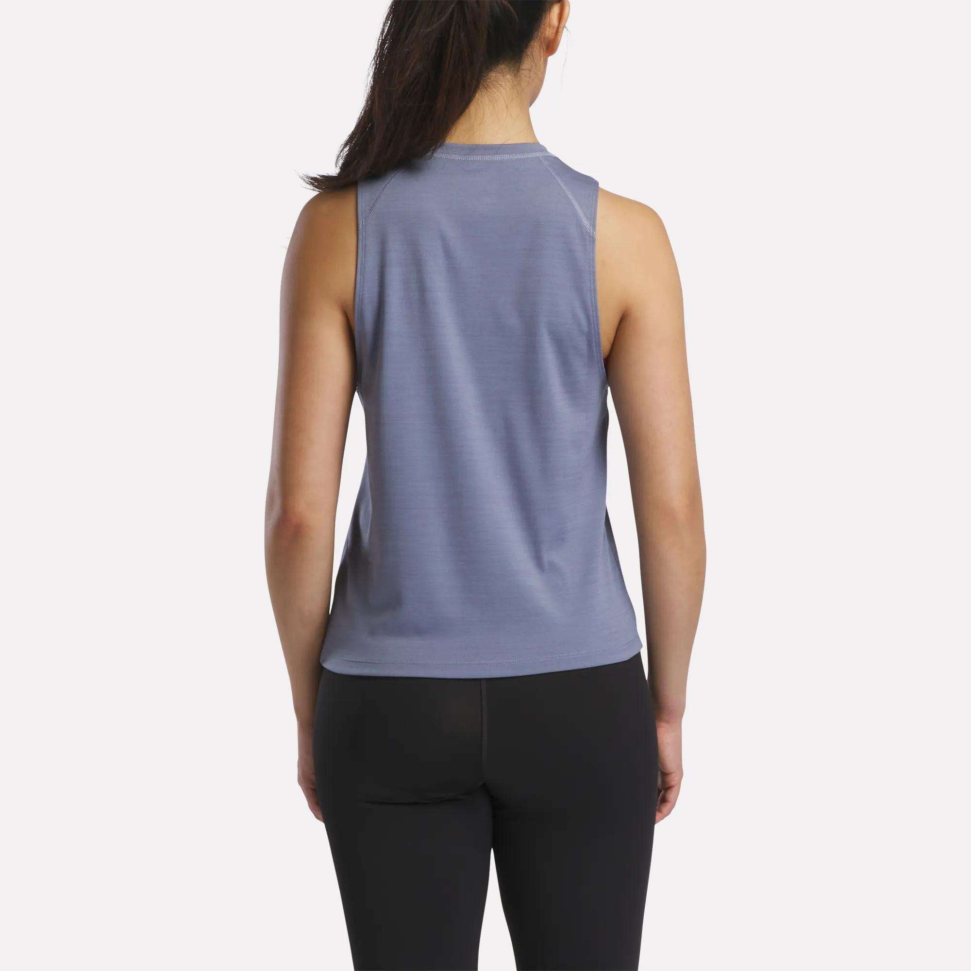 A woman with long dark hair in a ponytail is shown from behind, wearing a sleeveless blue Reebok Women's Training Tank Top and black leggings against a plain white background.