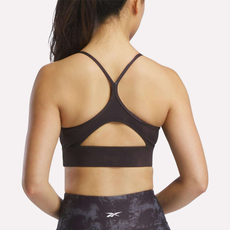 A woman with long hair in a ponytail is shown from the back, wearing the Reebok Women's Printed Tri-Back Sports Bra—a dark, sweat-wicking workout bra featuring thin straps and a keyhole cutout—paired with dark athletic leggings.