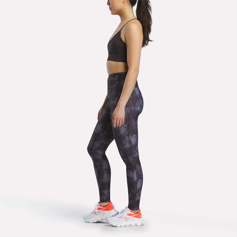 A woman wearing athletic apparel—including the Reebok Women's Printed Tri-Back Sports Bra, patterned leggings, and white sneakers—standing in profile against a plain white background.