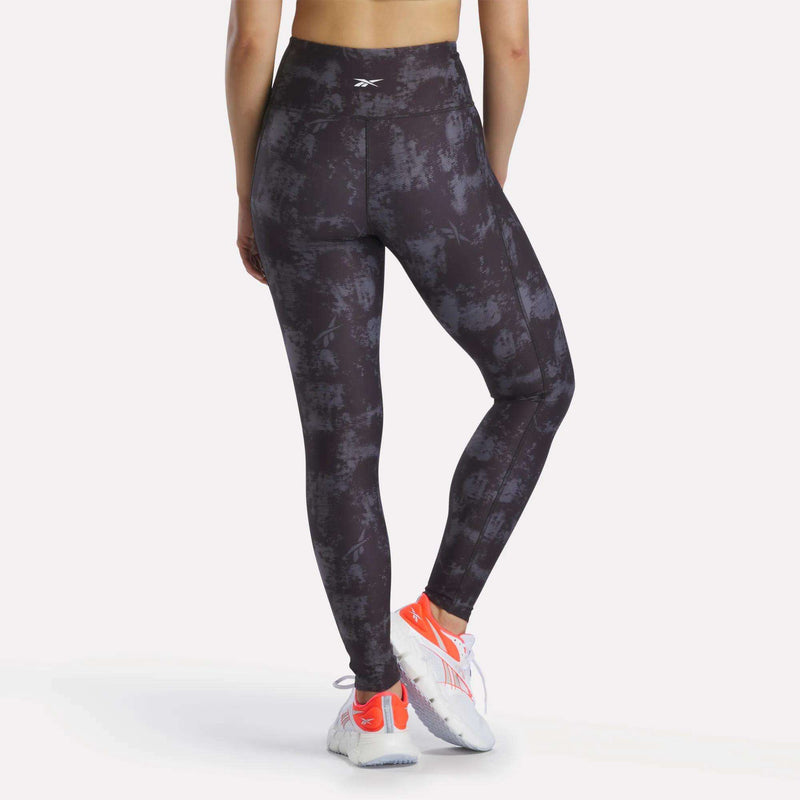 A person faces away, wearing Reebok Women's High-Rise Printed Leggings with sweat-wicking fabric, white and orange athletic shoes, and a sports top. The background is plain and light-colored.