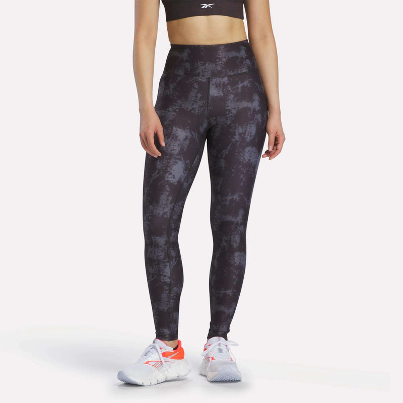 A woman in a black sports bra, Reebok Women's High-Rise Printed Leggings with sweat-wicking fabric, and white athletic shoes with orange accents stands against a plain light background.