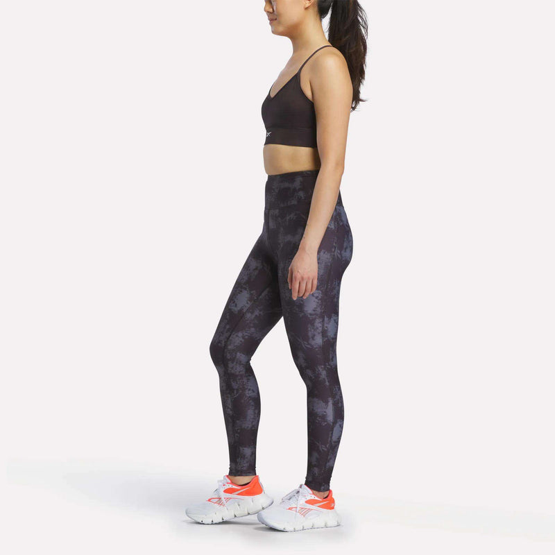 A woman stands sideways wearing Reebok Women's High-Rise Printed Leggings, a black sports bra, and white athletic shoes with orange details, against a plain white background.