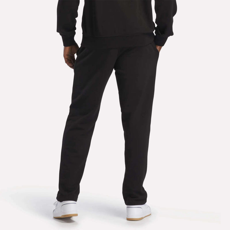 Wearing the Reebok Men's French Terry Jogger Pants, a person stands with hands in pockets, facing away from the camera against a light background. They're styled with a black sweatshirt and classic white sneakers for a comfortable, casual look.