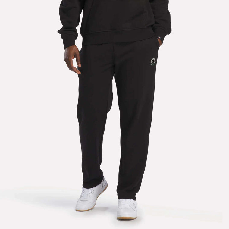 A person wearing Reebok Men's French Terry Jogger Pants in black, paired with a matching soft black sweatshirt and white Reebok sneakers, stands against a plain light background.