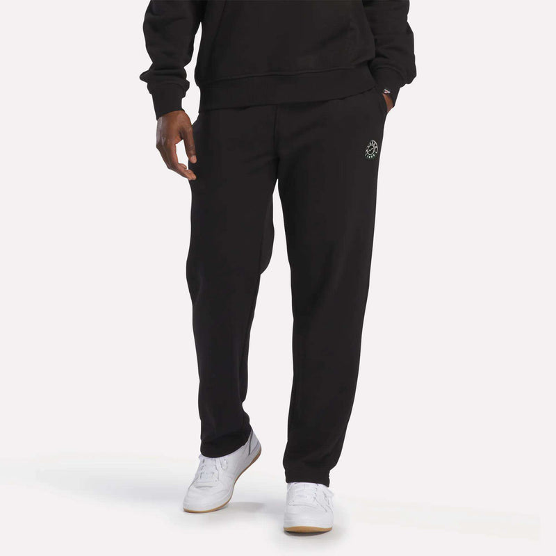 A person wearing Reebok Men's French Terry Jogger Pants in black, paired with a matching soft black sweatshirt and white Reebok sneakers, stands against a plain light background.