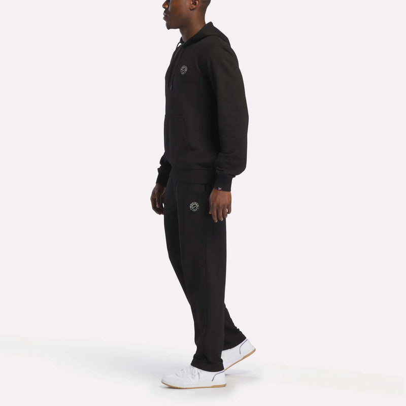 A man stands sideways wearing a matching black Reebok hoodie and the Men's French Terry Jogger Pants, paired with white sneakers. Both the hoodie and jogger pants feature a small circular Reebok logo and offer a soft feel against the skin. The background is plain white.