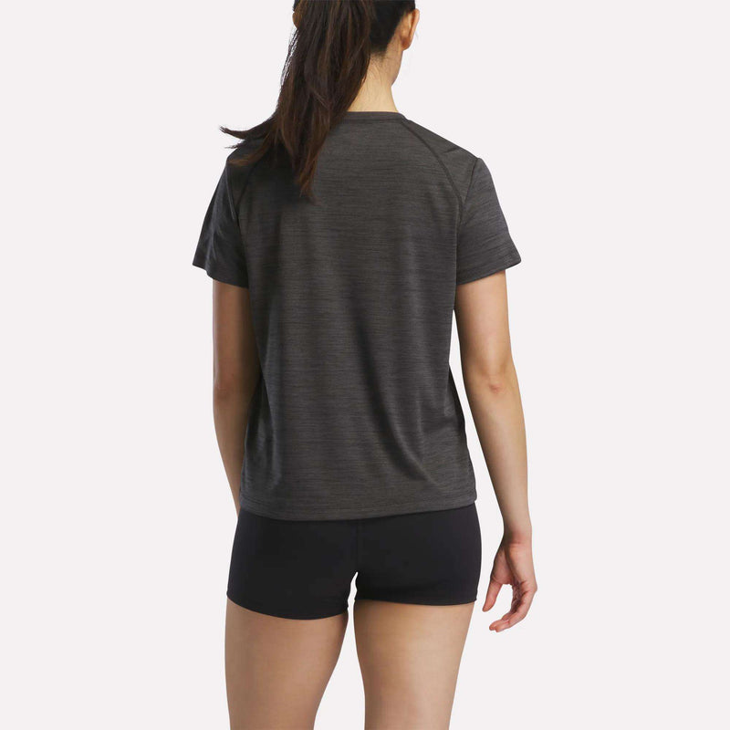 A woman with long brown hair in a ponytail stands with her back to the camera, wearing a dark gray Reebok Women's Training T-Shirt and black shorts against a plain light background.