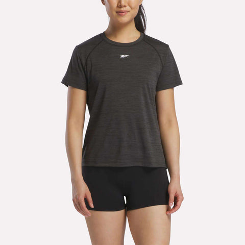 A woman stands against a plain background wearing a black Reebok Women's Training T-Shirt with a small white logo on the chest and black shorts.