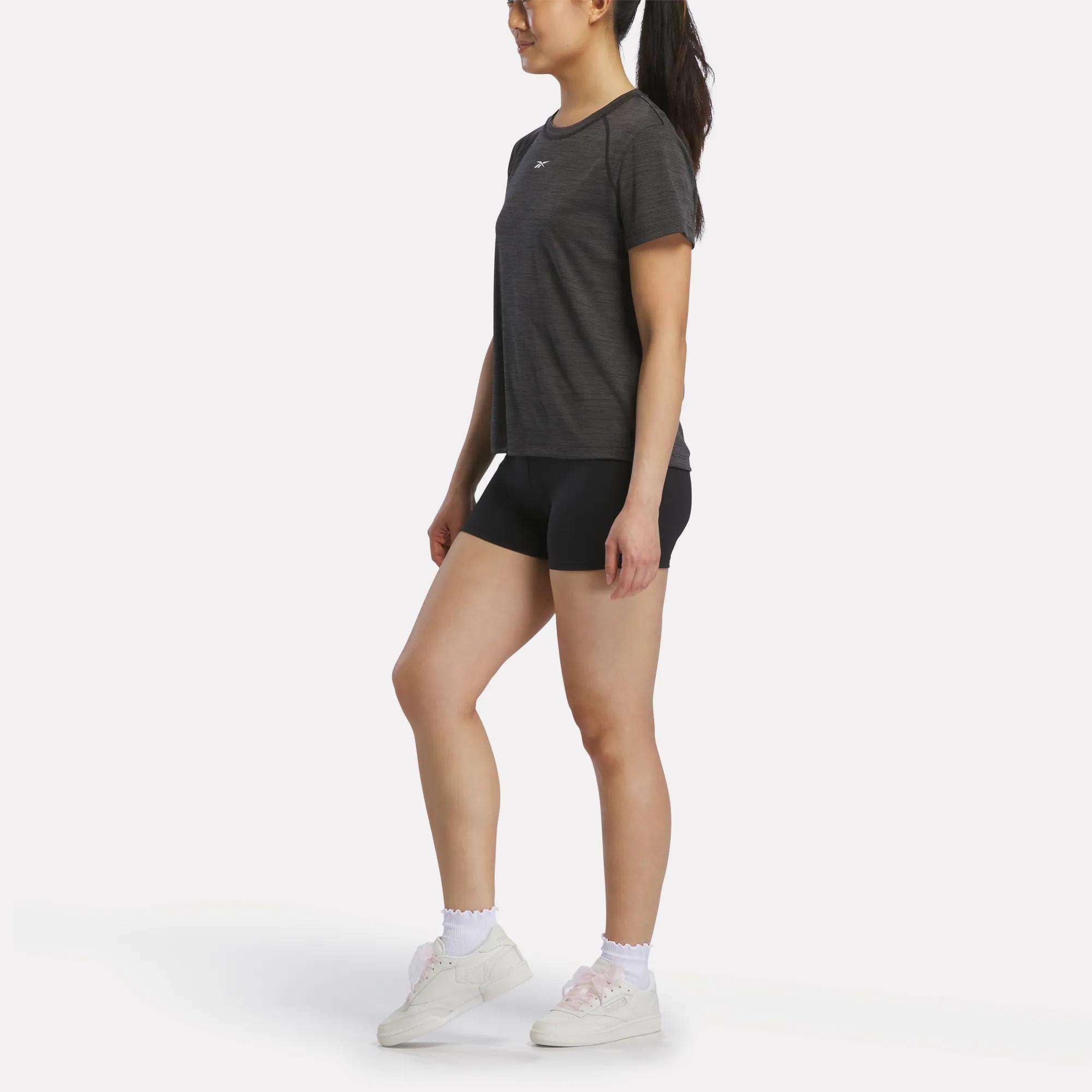 A woman with long dark hair tied back wears a Reebok Women's Training T-Shirt, black shorts, white socks, and white sneakers. She stands facing left against a plain white background.