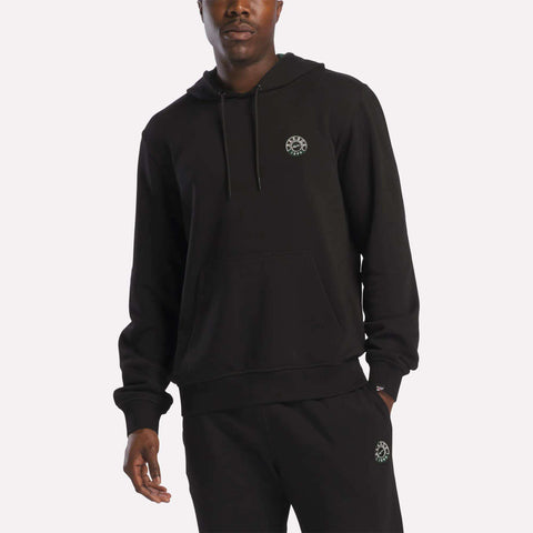A person wearing a black Reebok Men's French Terry Hoodie and matching black sweatpants, both featuring a small circular white emblem on the chest and thigh, stands against a plain light background.