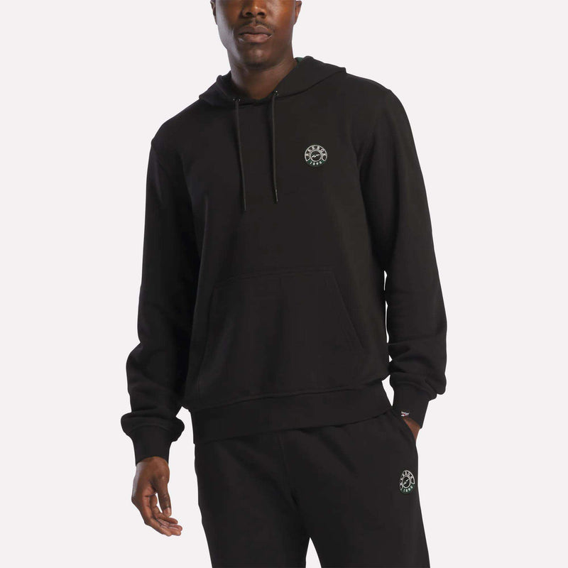 A person wearing a black Reebok Men's French Terry Hoodie and matching black sweatpants, both featuring a small circular white emblem on the chest and thigh, stands against a plain light background.