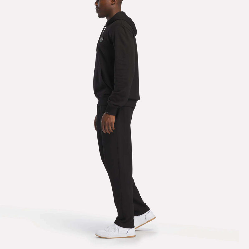 A person stands in profile wearing the Reebok Men's French Terry Hoodie, paired with black pants and white sneakers against a plain white background.