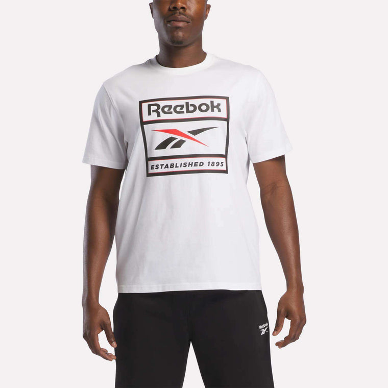 A man stands against a plain background wearing the Reebok Men's Logo T-Shirt in white, featuring the iconic Reebok logo and "Established 1895" printed on the front. Paired with black pants, this soft cotton tee from Reebok is ideal for everyday wear.