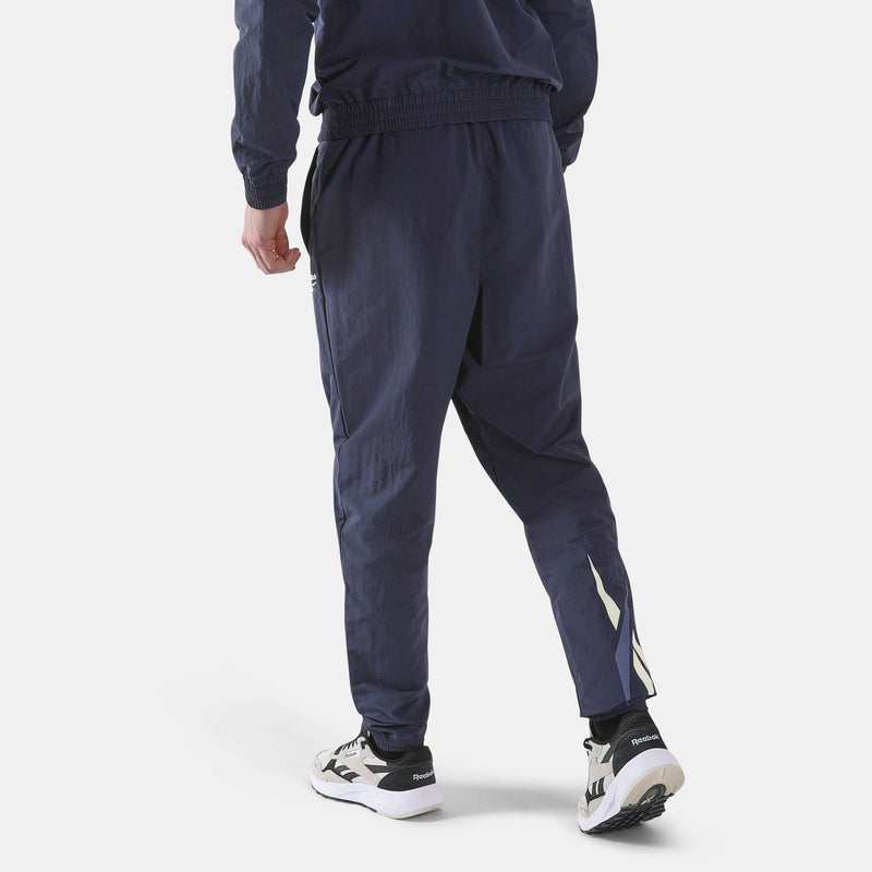A person viewed from behind wearing navy Men’s Classics Track Pants with a white and blue chevron graphic on the lower leg, paired with running shoes for casual comfort.