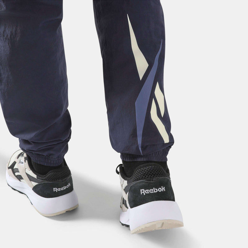 Close-up of navy blue Men’s Classics Track Pants with a large logo on the lower leg, worn with grey and black athletic sneakers for casual comfort.