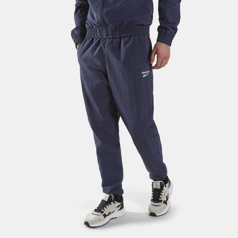 A person wearing dark blue Men’s Classics Track Pants with a small Reebok logo on the thigh and multicolored sneakers.