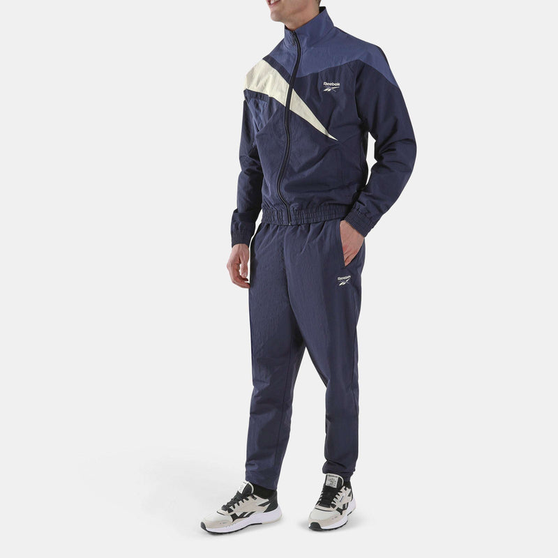 A person wearing a navy, blue, and cream color-blocked Reebok track jacket with matching Men’s Classics Track Pants and athletic shoes for casual comfort.