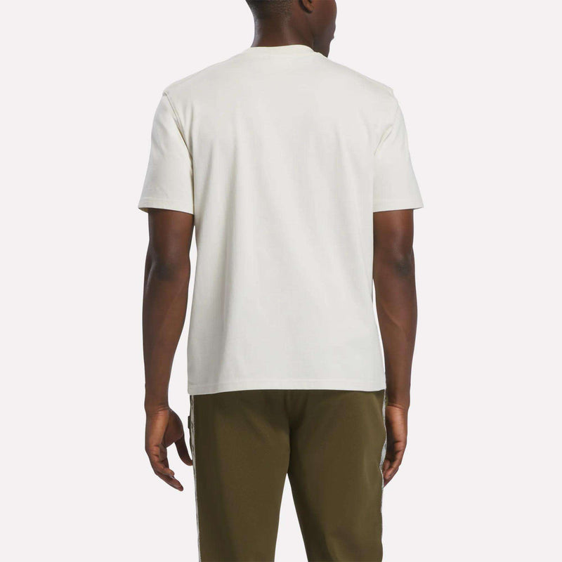 A person is standing with their back to the camera, wearing a plain white Reebok Men’s Heritage T-Shirt made from soft cotton jersey and olive green pants against a light background.