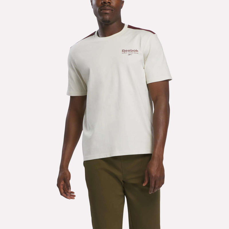 A man wearing a comfortable Reebok Men’s Heritage T-Shirt in linen, crafted from soft cotton jersey, stands against a plain white background. The shirt features brown shoulder accents and is paired with olive green pants.