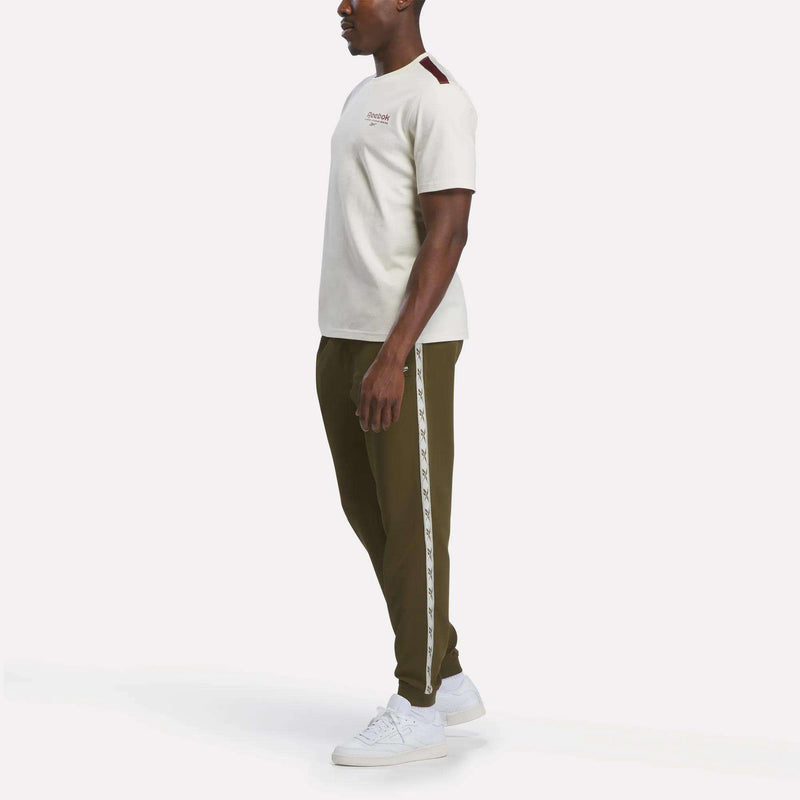 A man stands in profile wearing the Reebok Men’s Heritage T-Shirt, made from soft cotton jersey for ultimate comfort. He pairs it with olive green jogger pants featuring a light-colored side stripe and white sneakers, all set against a plain white background.