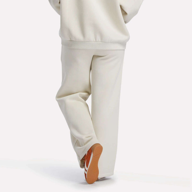 A person in off-white Reebok Women's Heritage Flared Pants and a matching sweatshirt stands with one leg crossed behind the other, revealing the sole of their orange and white sneakers against a plain white background.