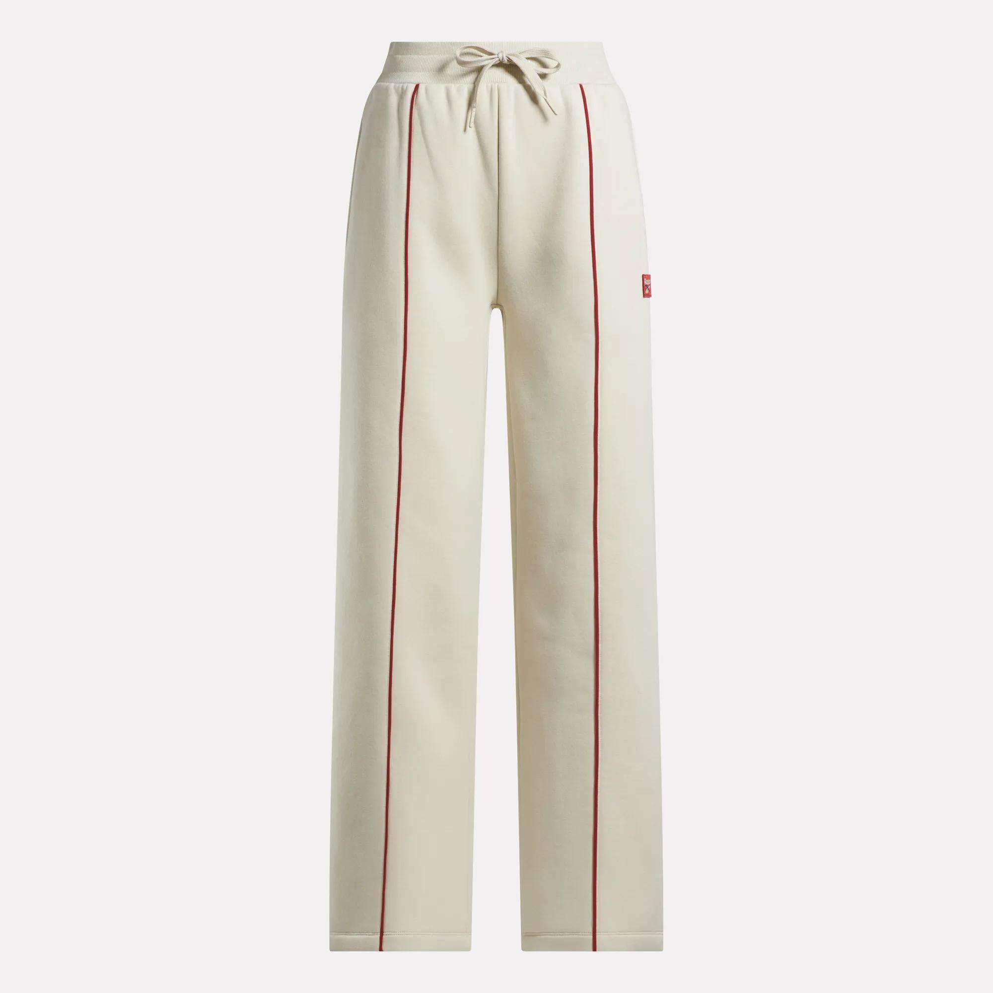 The Reebok Women's Heritage Flared Pants are beige sweatpants made from soft brushed fleece, featuring red piping on the front seams, an elastic drawstring waistband, and side pockets.