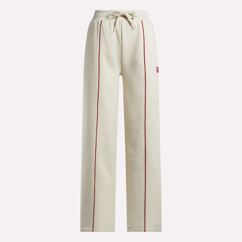 The Reebok Women's Heritage Flared Pants are beige sweatpants made from soft brushed fleece, featuring red piping on the front seams, an elastic drawstring waistband, and side pockets.