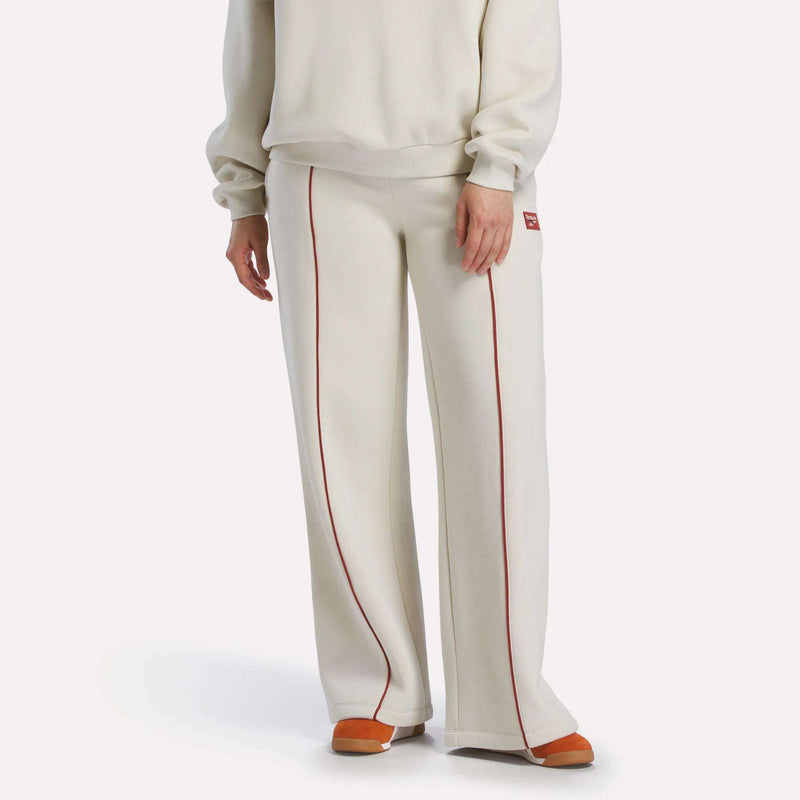 A person wears Reebok Women's Heritage Flared Pants in off-white with red piping, paired with an off-white sweater and orange-beige slippers, creating a sporty vibe against a plain white background.