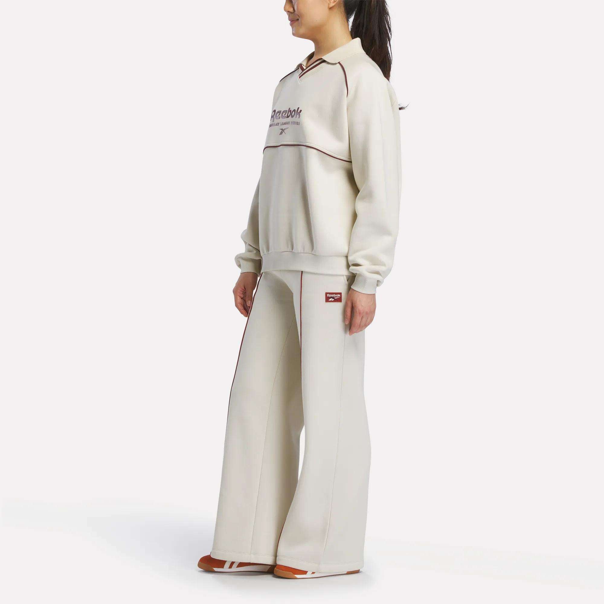 A woman poses sideways in a cream Reebok outfit, featuring the Women's Heritage Flared Pants and matching sweatshirt with chest text and logo, paired with brown shoes. The scene is set against a plain white background.