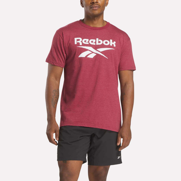 A person models Reebok's Men's Stacked Vector T-Shirt in maroon with a bold white logo, paired with black Reebok shorts, against a plain white background.