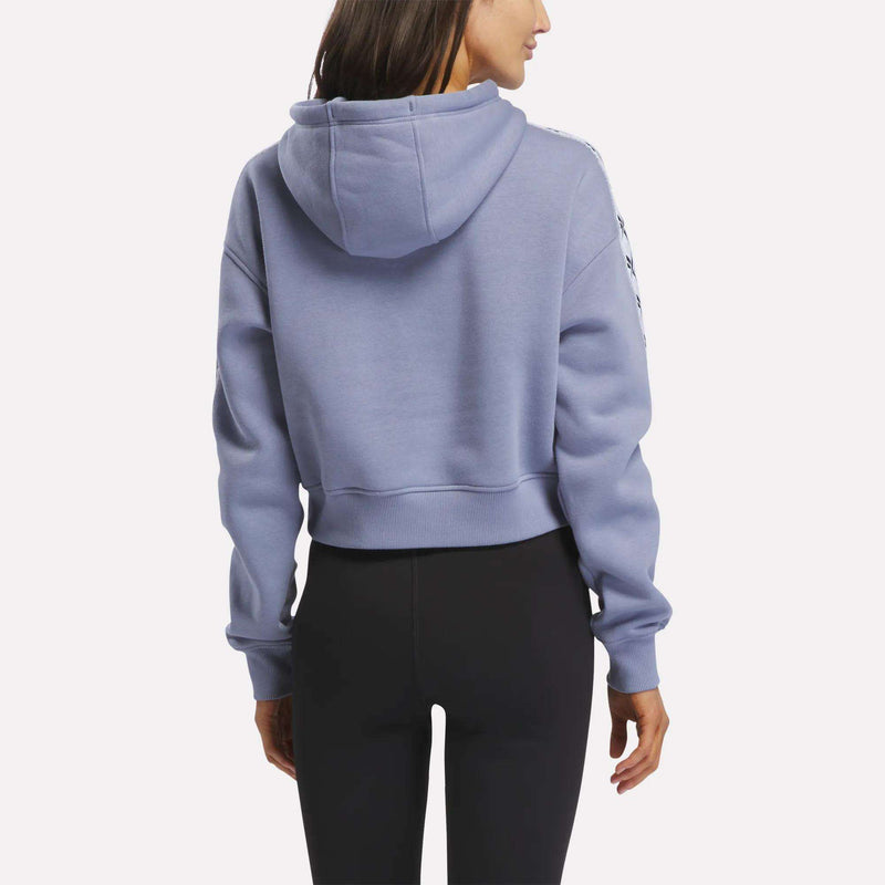 A woman is shown from the back, with her head turned slightly to the side, wearing a light purple Reebok Women’s Cropped Heritage Hoodie and black leggings.