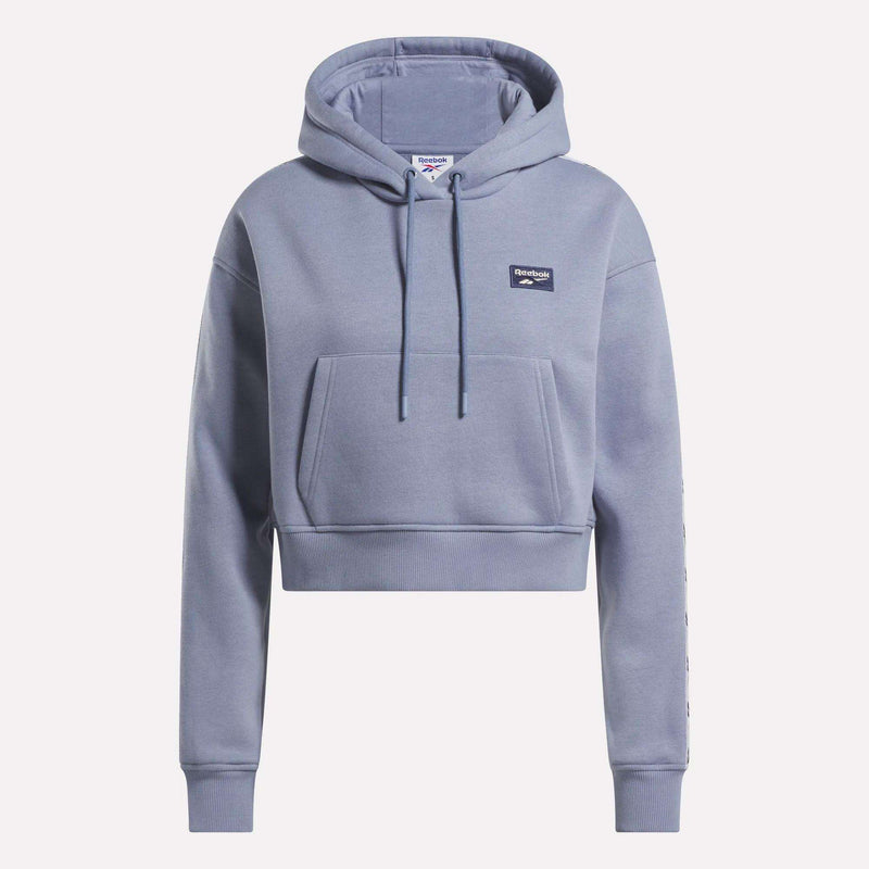 Introducing the Reebok Women’s Cropped Heritage Hoodie—a stylish light blue piece featuring long sleeves, a front pocket, and a drawstring hood. The soft and comfortable fabric is complemented by a small rectangular logo on the chest, making it the perfect addition to your casual wardrobe.