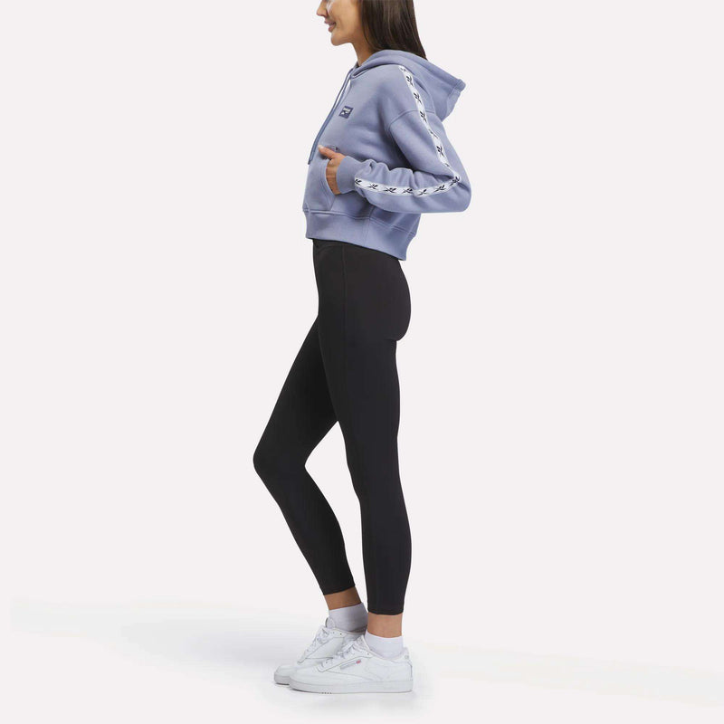 A woman stands sideways against a plain white background, wearing the Reebok Women’s Cropped Heritage Hoodie in light blue with patterned sleeves, paired with black leggings and white sneakers.