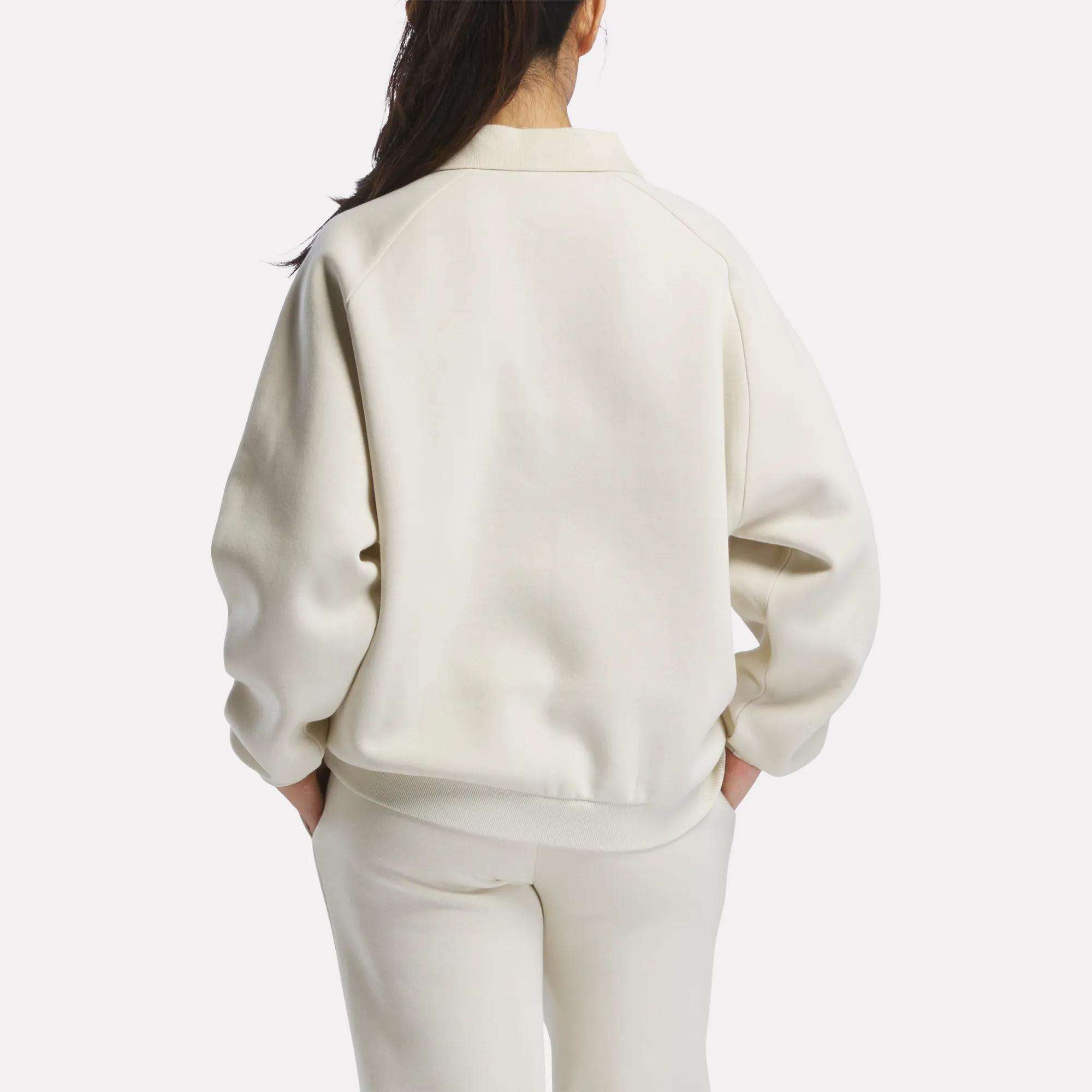A woman with long dark hair, wearing a cream Women's Heritage Crewneck Sweatshirt by Reebok and matching pants with subtle branding, stands facing away against a plain white background.