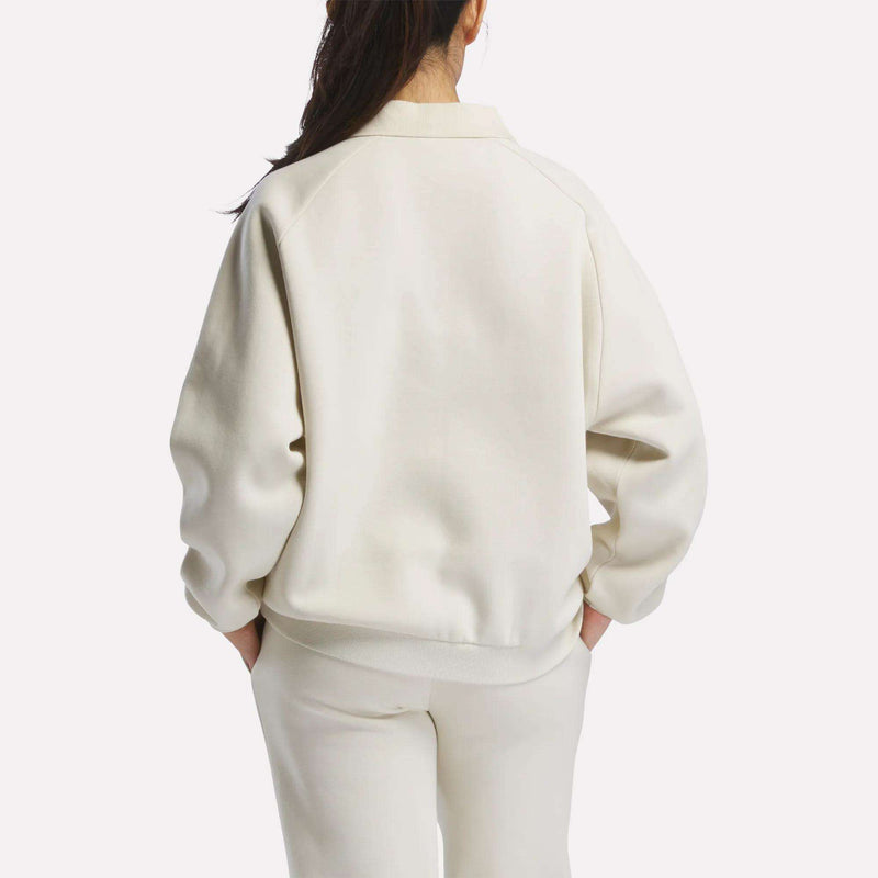 A woman with long dark hair, wearing a cream Women's Heritage Crewneck Sweatshirt by Reebok and matching pants with subtle branding, stands facing away against a plain white background.