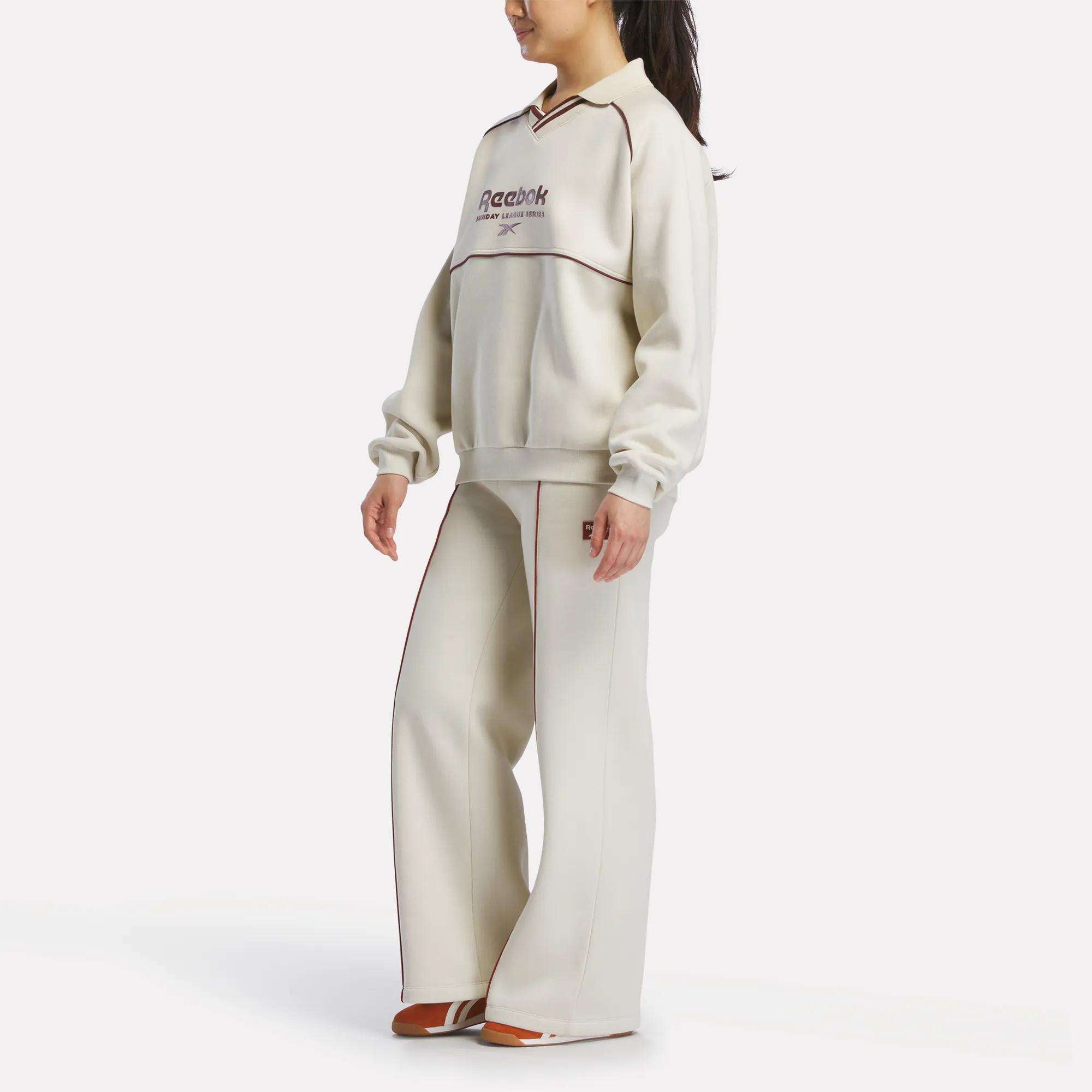 A woman with her hair in a ponytail wears the Reebok Women's Heritage Crewneck Sweatshirt, featuring cream color, logo detail, and wide-leg pants with brown trim, paired with brown shoes and standing slightly to the side.