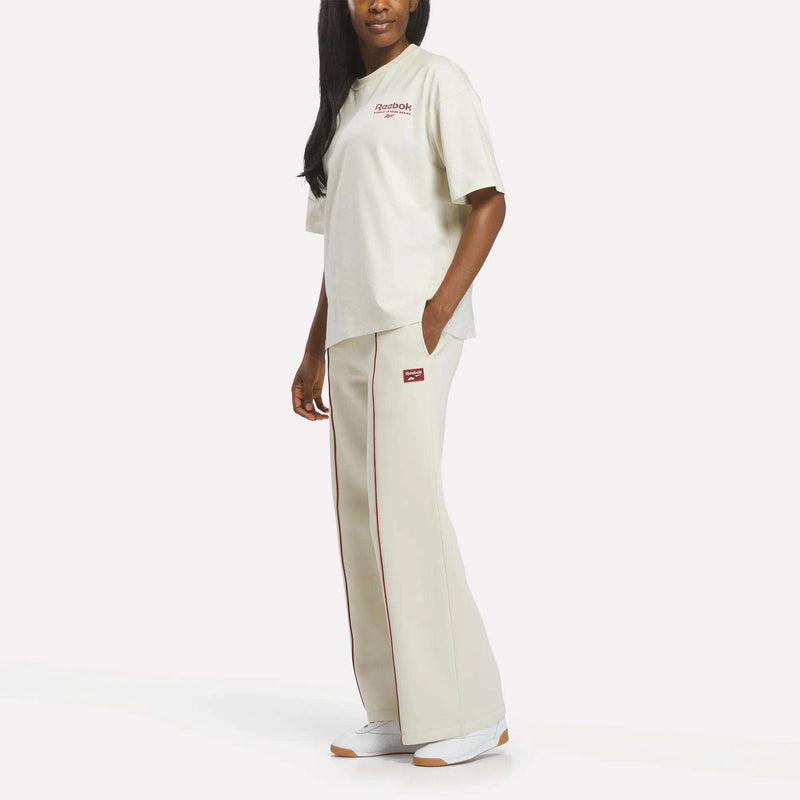 A person stands against a plain white background wearing a cream Women's League Club Graphic T-Shirt by Reebok with matching wide-leg pants featuring red trim and white sneakers.