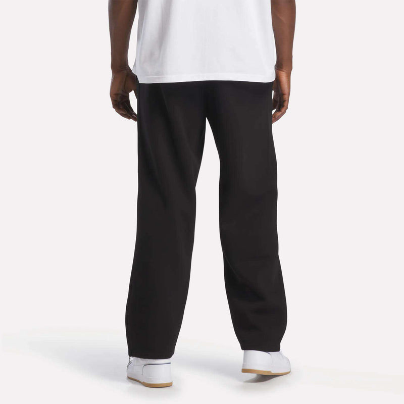 A person is standing facing away, wearing a white T-shirt, Men’s International Straight Leg Pants by Reebok in plush fleece with a relaxed open hem, and white Reebok sneakers. The background is plain and well-lit, showing only the lower part of their body.