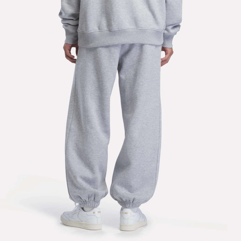 A person stands facing away, wearing light gray Reebok Men’s Relaxed Logo Joggers with elastic cuffs, a matching fleece sweatshirt, and white Reebok sneakers.