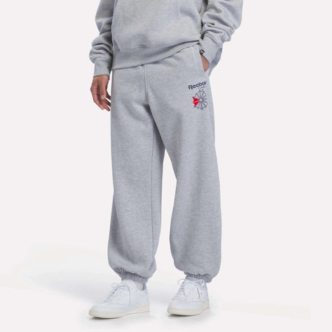 A person is shown from the waist down wearing Reebok Men’s Relaxed Logo Joggers in Light Grey Marl, paired with white sneakers, standing against a plain white background. Their hands are tucked into the jogger pockets.