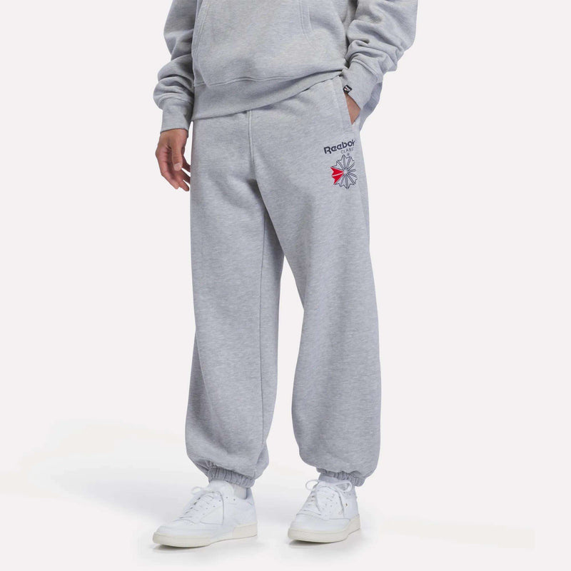 A person is shown from the waist down wearing Reebok Men’s Relaxed Logo Joggers in Light Grey Marl, paired with white sneakers, standing against a plain white background. Their hands are tucked into the jogger pockets.