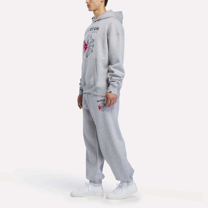 A person stands against a plain white background wearing Men’s Relaxed Logo Joggers and a gray Reebok fleece hoodie, both featuring red and black Reebok logos, paired with white sneakers.