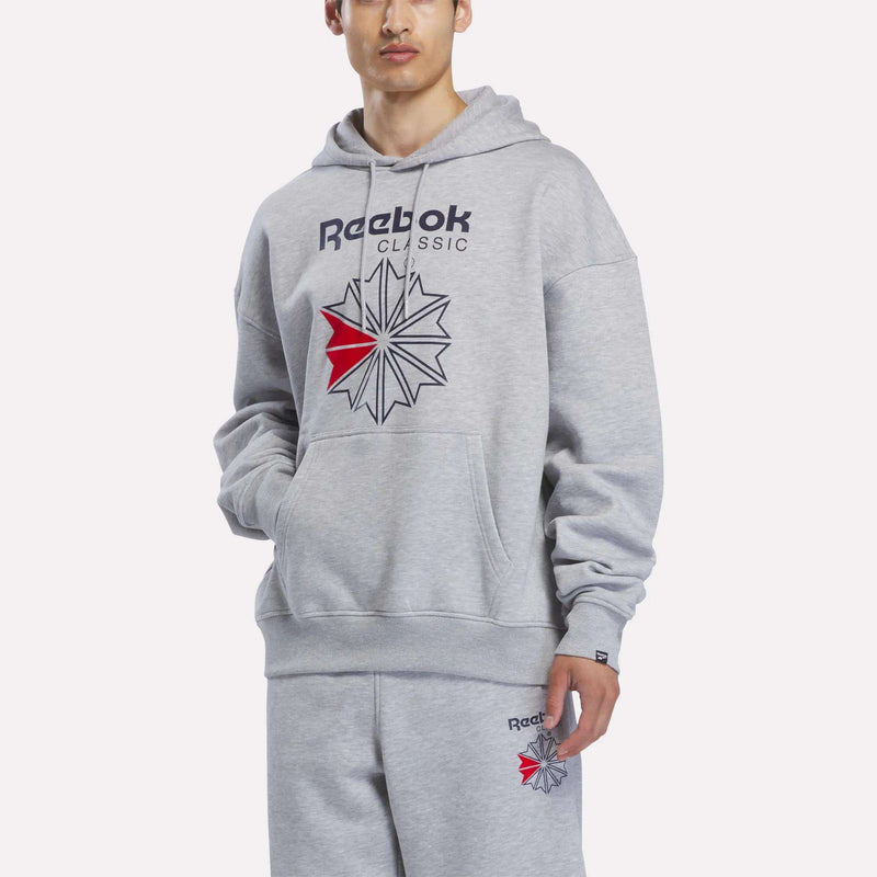 A man stands against a plain white background wearing the Reebok Men’s Starcrest Relaxed Hoodie and matching light grey marl sweatpants, both featuring a bold red and black Starcrest logo. Made from soft fleece, this classic hoodie offers ultimate comfort.