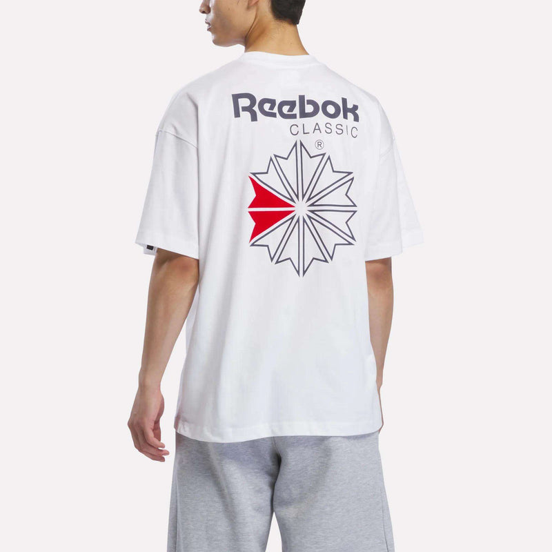A person wearing the Reebok Men’s Relaxed Logo T-Shirt, crafted from soft cotton jersey and designed with a bold star logo and red detail on the back, paired with light gray shorts, stands against a plain background.