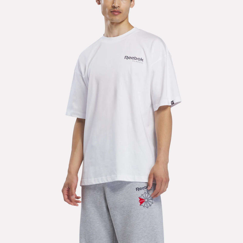 A person wearing a white Reebok Men’s Relaxed Logo T-Shirt and light gray shorts stands against a plain background, facing slightly left.