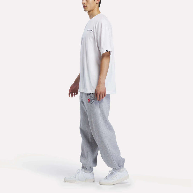 A person stands sideways against a plain white background wearing the Reebok Men’s Relaxed Logo T-Shirt, crafted from soft cotton jersey, paired with light gray graphic sweatpants and white sneakers.