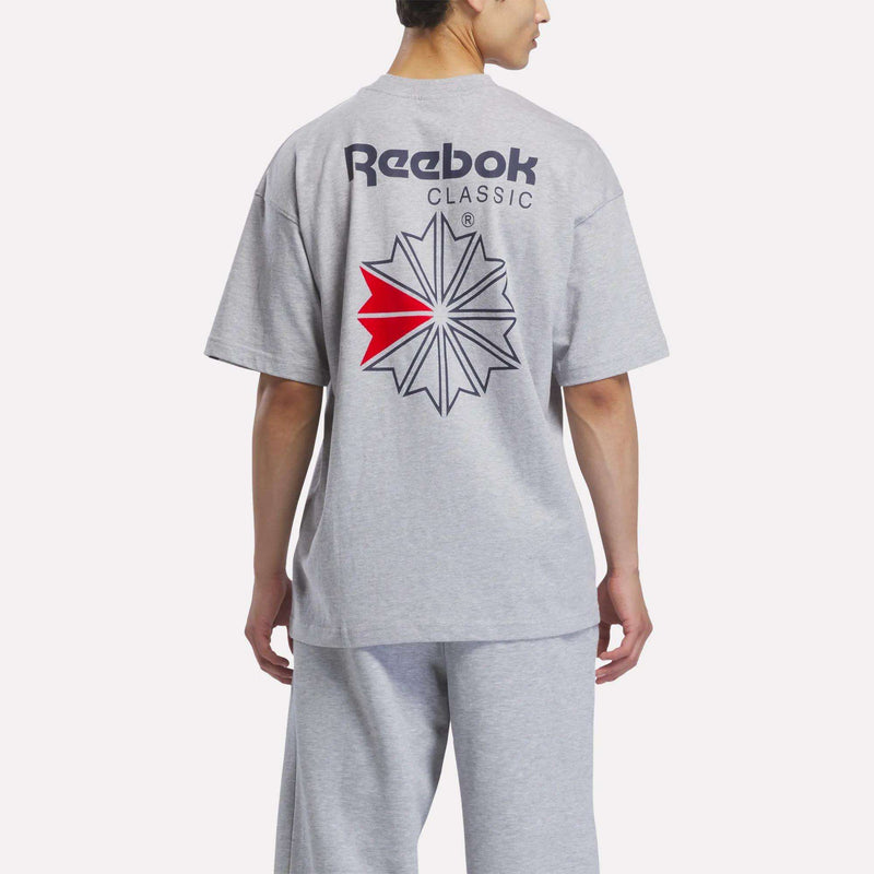 A man is shown from the back wearing a light gray Reebok Men’s Relaxed Logo T-Shirt and pants. This cotton tee features a large black, red, and white Reebok logo graphic on the back.